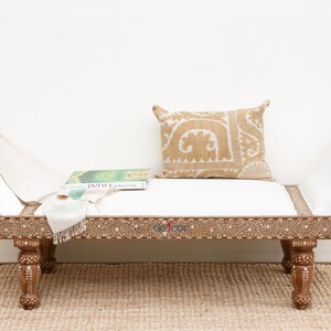 May include: A white upholstered bench with a carved wooden frame and legs. The bench has a white cushion and a tan patterned pillow. A book titled "India" is resting on the cushion.