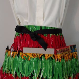 以下が含まれることがあります： 赤と緑の唐辛子のプリントが施されたエプロンで、黒いリボンで結ばれています。エプロンには複数のポケットと、右側に大きなポケットがあります。