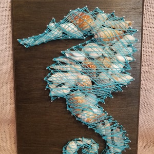 May include: A teal blue string art piece depicting a seahorse on a dark brown wooden background. The seahorse is made of string wrapped around nails and is decorated with seashells.