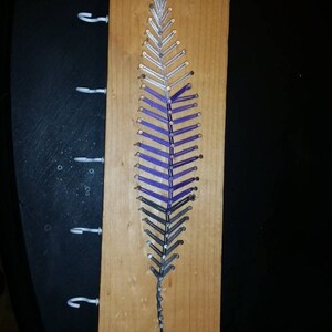 Puede incluir: Un porta llaves de madera con tres ganchos y un diseño de arte de hilo de una pluma en tonos de morado, gris y plateado.