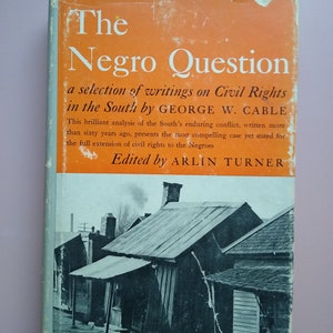 以下が含まれることがあります： 「The Negro Question: A Selection of Writings on Civil Rights in the South by George W. Cable」というタイトルの本の表紙。本の表紙はオレンジ色で、白い文字が書かれている。表紙の下部には、小さな町の通りの白黒写真が写っている。