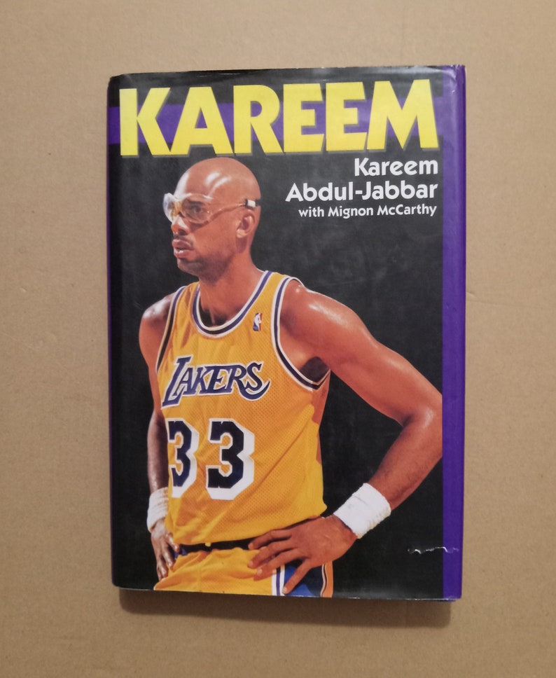 May include: A book cover featuring a photo of Kareem Abdul-Jabbar wearing a yellow Lakers jersey with the number 33. The title of the book is "Kareem" and the author is Kareem Abdul-Jabbar with Mignon McCarthy.