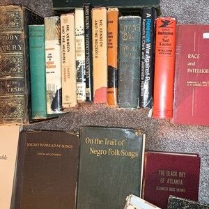 Puede incluir: Una colecci&oacute;n de libros antiguos con t&iacute;tulos relacionados con la raza, la esclavitud y la experiencia negra en Am&eacute;rica. Los libros est&aacute;n dispuestos en una fila sobre una superficie marr&oacute;n claro.