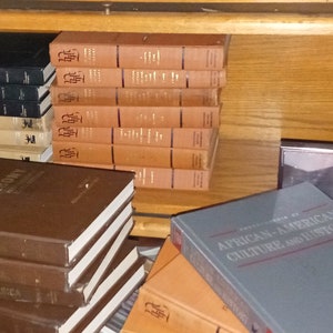 May include: A wooden bookshelf filled with books. The books are stacked in rows and are mostly brown and black. Some of the books have titles visible, such as "Ebony" and "African-American Culture and History".