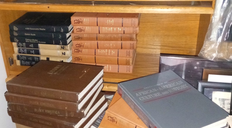 May include: A wooden bookshelf filled with books. The books are stacked in rows and are mostly brown and black. Some of the books have titles visible, such as "Ebony" and "African-American Culture and History".