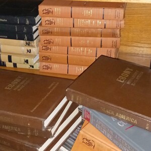 May include: A stack of brown hardcover books with the title "EBONY" and "BLACK AMERICA" visible. The books are on a wooden shelf with other books in the background.