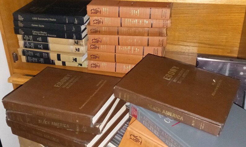 May include: A stack of brown hardcover books with the title "EBONY" and "BLACK AMERICA" visible. The books are on a wooden shelf with other books in the background.