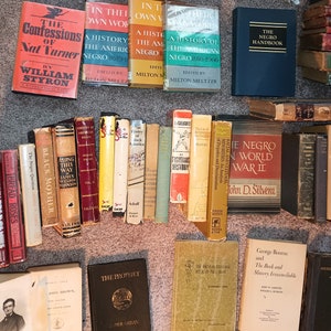 Puede incluir: Una colecci&oacute;n de libros antiguos de tapa dura sobre la historia y la cultura afroamericana. Los libros est&aacute;n dispuestos en una fila sobre una alfombra beige. Algunos de los t&iacute;tulos incluyen "The Negro World War II", "The Negro Question", "Black Mother", "Along This Way", "The Prophet", "The American Negro 1619-1966", y "In Their Own Words".