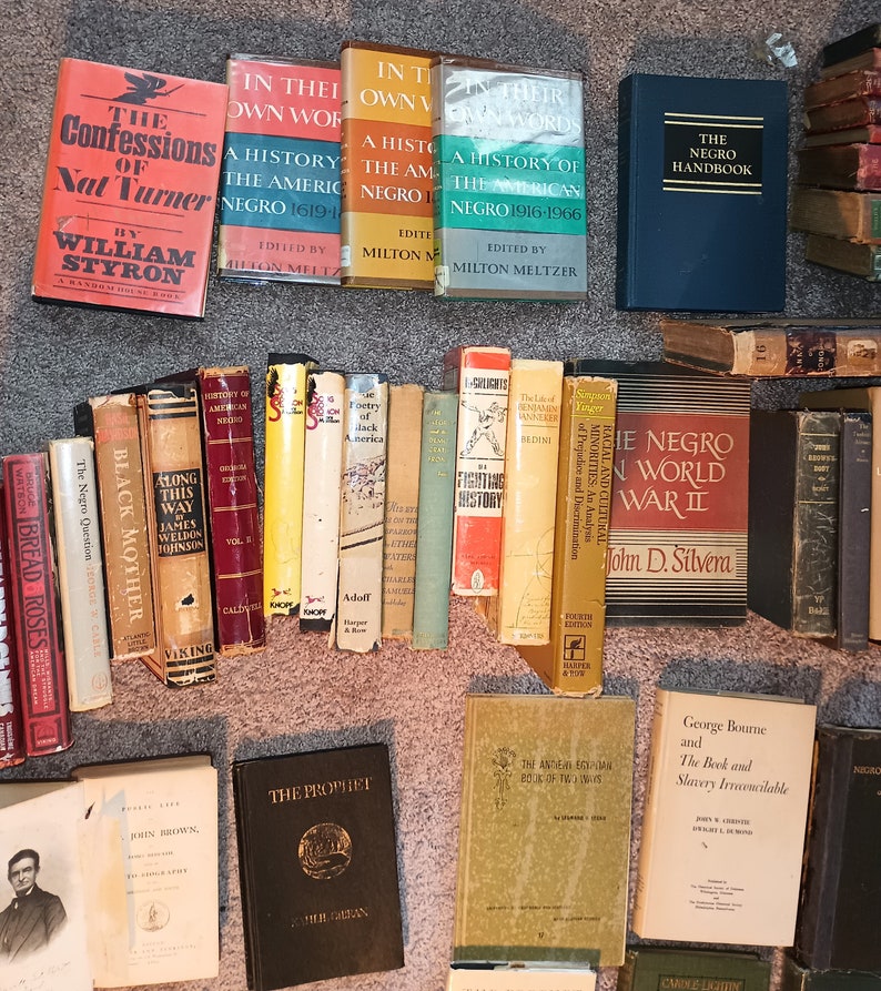 Puede incluir: Una colecci&oacute;n de libros antiguos de tapa dura sobre la historia y la cultura afroamericana. Los libros est&aacute;n dispuestos en una fila sobre una alfombra beige. Algunos de los t&iacute;tulos incluyen "The Negro World War II", "The Negro Question", "Black Mother", "Along This Way", "The Prophet", "The American Negro 1619-1966", y "In Their Own Words".