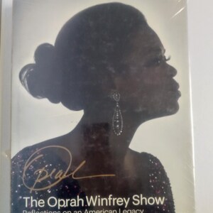 May include: A black and white silhouette of Oprah Winfrey with a gold signature and the text "The Oprah Winfrey Show Reflections on an American Legacy".