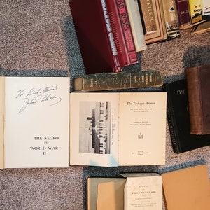 Puede incluir: Una colecci&oacute;n de libros antiguos con varios t&iacute;tulos, incluyendo "Bread Roses", "Black Mother", "The Negro in World War II" y "The Tuskegee Airmen". Los libros est&aacute;n dispuestos sobre una alfombra marr&oacute;n.