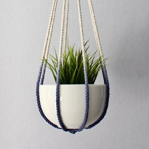 Op de afbeelding: Een witte keramische plantenpot met een groene plant die hangt aan een macrame plantenhanger met een blauwe kwast.
