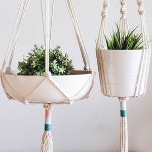 May include: Two macrame plant hangers with white cord and colourful accents. The larger hanger has a white ceramic pot with a green plant. The smaller hanger has a white ceramic pot with a green plant. Both hangers have a white tassel.