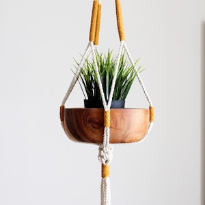 May include: A white macrame plant hanger with yellow accents holds a wooden planter with a green plant. The hanger has a tassel at the bottom.