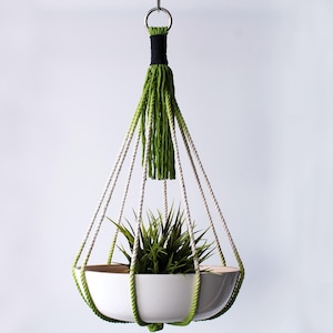 May include: A white ceramic hanging planter with a green plant inside. The planter is suspended by a macrame rope with a green and white tassel.