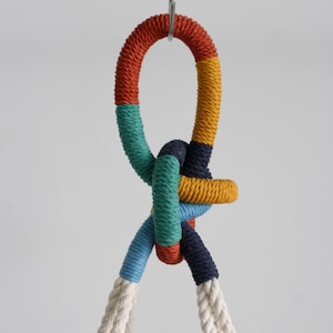 Peut inclure: Gros plan sur un nœud macramé coloré. La corde est enveloppée dans des sections rouges, turquoise, jaunes, bleues et marine. Des brins de corde blanc cassé pendent en dessous. Le nœud est suspendu à un crochet en métal.
