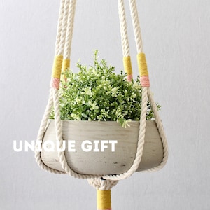 May include: A macrame plant hanger with a concrete pot and a green plant. The hanger has yellow and pink accents. The text "UNIQUE GIFT" is visible in the image.