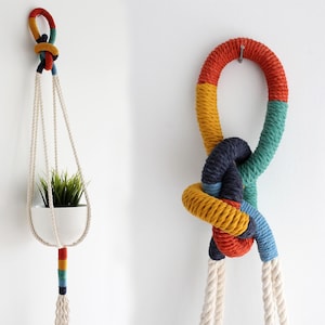 Puede incluir: Dos colgadores de plantas de macramé. Uno sostiene una maceta de cerámica blanca con vegetación. Los colgadores están hechos de cuerda color crema con detalles coloridos en rojo, amarillo, verde y azul. El segundo es un nudo decorativo.