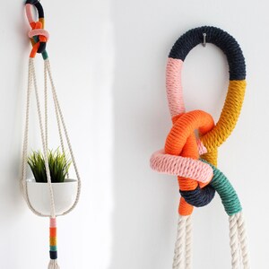 Pode incluir: Duas suspensões de plantas em macramé. Uma segura um vaso de cerâmica branco com vegetação. A outra é um nó decorativo. Ambas são feitas de corda colorida em tons de rosa, laranja, amarelo, verde e azul, com detalhes em corda branca.