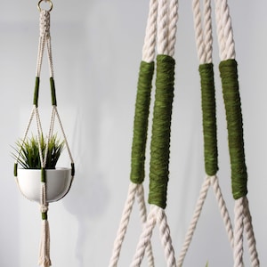 Peut inclure: Un porte-plante en macramé blanc avec des accents verts. Le porte-plante est fait de corde de coton et a un bol au fond pour contenir une plante. Le porte-plante est suspendu à un mur blanc.