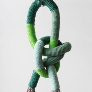 Peut inclure: Un porte-plante en macramé avec un sommet noué en corde verte et grise. Le porte-plante est accroché à un crochet en métal.