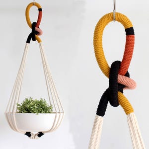 May include: Two hanging planters with a white ceramic pot and a plant. The planters are suspended by cream-coloured rope and colourful knotted accents in yellow, red, black, and pink. The background is a plain white wall.