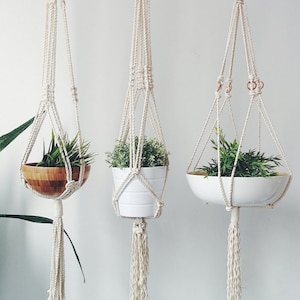 May include: Three macrame plant hangers with different styles of pots. The hangers are made of natural white cotton rope and have tassels at the bottom. The pots are a wooden bowl, a white ceramic pot, and a white ceramic bowl.