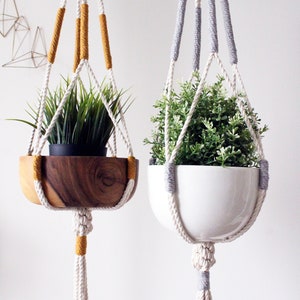 Peut inclure: Deux suspensions macramé pour plantes avec des pots en bois et en céramique blanche. La suspension de gauche a un cordon jaune et blanc et la suspension de droite a un cordon gris et blanc.