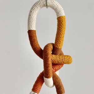 May include: A hanging macrame plant holder with a knotted design. The rope is white, mustard yellow, and brown. The top loop is suspended from a metal hook. The design is a modern take on a classic craft.
