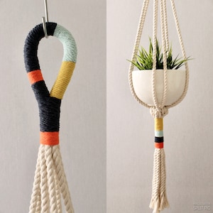 May include: A macrame plant hanger with a white ceramic pot and green plant. The hanger is made of natural coloured rope with black, orange, yellow, and light blue accents.