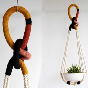 May include: Two hanging plant holders made of thick rope. The left holder features a large knot with sections of mustard yellow, terracotta, and black. The right holder has a similar knotted top and holds a white bowl containing a green plant.
