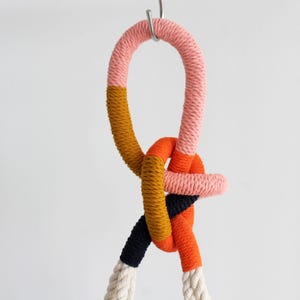 Könnte beinhalten: Dekorative, geknotete Seilskulptur in verschiedenen Farben. Das Seil hat Abschnitte in Rosa, Orange, Senfgelb, Marineblau und Weiß. Das Seil ist geschlungen und geknotet und hängt an einem Metallhaken vor einem schlichten weißen Hintergrund.