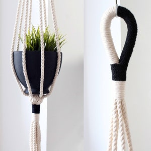 May include: Two macrame plant hangers with black accents. The first hanger is holding a black pot with a green plant. The second hanger is hanging from a hook and has a black accent at the top.