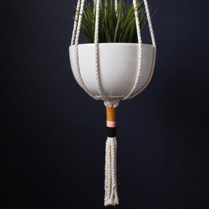 May include: White ceramic planter with green plants hanging from a macrame plant hanger with a white tassel and pink, black, and gold accents.