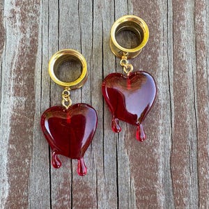 May include: A pair of gold-colored ear gauges with dangling red heart-shaped charms. The hearts have a dripping effect, giving them a unique and eye-catching appearance. The gauges are set against a rustic wooden background.
