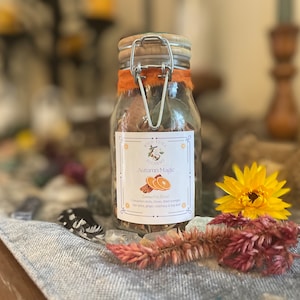 Op de afbeelding: Een glazen pot met een houten deksel en metalen sluiting, gevuld met potpourri. De pot heeft een label met de tekst "Autumn Magic" met afbeeldingen van sinaasappels en kaneelstokjes. De pot is versierd met oranje stof.