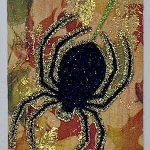 Könnte beinhalten: Eine schwarze Spinne mit goldenen Glitzerakzenten auf einem gemusterten Hintergrund. Der Text "it's all perspective" ist auf einem weißen Streifen oben im Bild gedruckt.