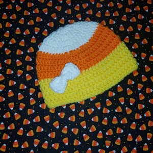 May include: A crocheted baby hat shaped like a candy corn with white, orange, and yellow stripes. The hat has a white crocheted bow on the front.