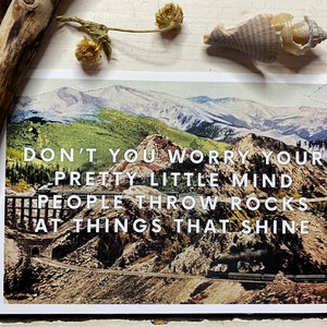 May include: A vintage postcard with a scenic mountain landscape featuring a train track and a train in the distance. The postcard has the text "Don't you worry your pretty little mind people throw rocks at things that shine."