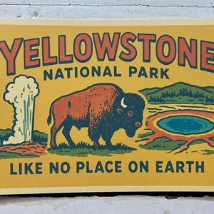 May include: A rectangular print featuring a yellow background with the text YELLOWSTONE NATIONAL PARK at the top and LIKE NO PLACE ON EARTH at the bottom. The illustration depicts a brown bison, a white geyser, and a blue thermal pool.