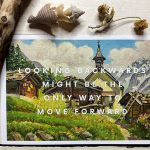 May include: A rectangular postcard featuring an oil painting of a mountain village with wooden cabins, a church, and green fields. The white text overlay reads: LOOKING BACKWARDS MIGHT BE THE ONLY WAY TO MOVE FORWARD. The print is displayed on a light surface.