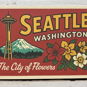 May include: A rectangular print featuring a red background with the text SEATTLE WASHINGTON and The City of Flowers. The design includes a yellow Space Needle, a green mountain with a snow-capped peak, and a cluster of red, yellow, and white flowers.