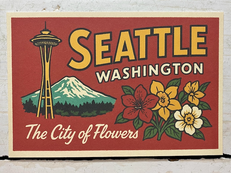 May include: A rectangular print featuring a red background with the text SEATTLE WASHINGTON and The City of Flowers. The design includes a yellow Space Needle, a green mountain with a snow-capped peak, and a cluster of red, yellow, and white flowers.