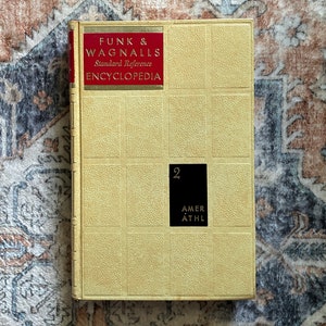 May include: A yellow hardcover Funk & Wagnalls Standard Reference Encyclopedia volume 2 book. The cover features a red rectangular label with gold text and a black rectangular label with the number 2 and the text AMER ATHL in gold.
