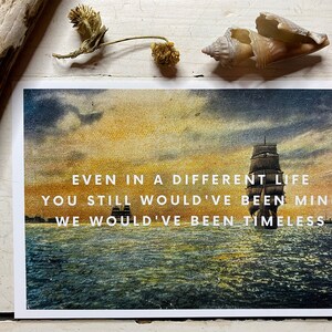 May include: A rectangular print featuring a sunset seascape with a large sailing ship and the white text: EVEN IN A DIFFERENT LIFE YOU STILL WOULD'VE BEEN MINE WE WOULD'VE BEEN TIMELESS. The image displays deep blue water and a golden sky.