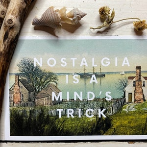 May include: A rectangular postcard featuring a vintage-style landscape illustration of coastal houses and a sailing ship on the water. The white text overlay reads NOSTALGIA IS A MIND'S TRICK. The print is surrounded by two pieces of driftwood, a seashell, and a dried flower.