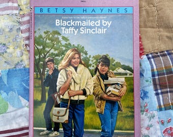 May include: A paperback book titled Blackmailed by Taffy Sinclair by Betsy Haynes. The cover features an illustration of three young people outdoors, with one person holding a stack of books and another wearing a cream-colored jacket and carrying a bag.