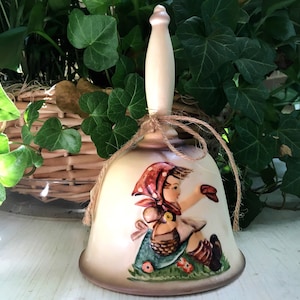 May include: A ceramic bell with a white handle and a painted image of a girl in a red and white dress holding a basket. The bell is tied with a brown twine.