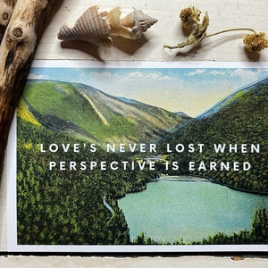 May include: A rectangular postcard featuring a landscape illustration of a lake surrounded by dense green forests and mountains. The text LOVE'S NEVER LOST WHEN PERSPECTIVE IS EARNED is printed in white capital letters across the center of the image.