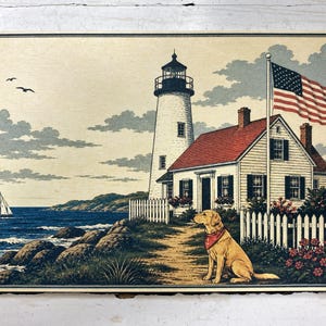May include: A rectangular print featuring a coastal scene with a white lighthouse, a house with a red roof, and a yellow dog wearing a red bandana sitting on a path. A sailboat floats on the blue ocean, and an American flag flies on a pole.